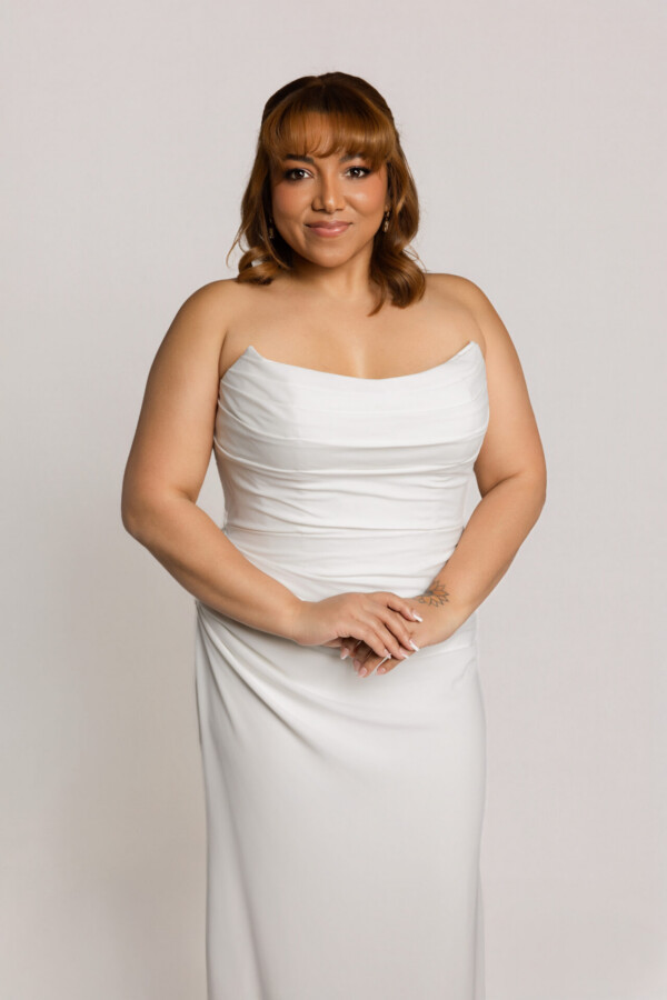 Close-up of a bride wearing a draped one-shoulder white wedding gown with a fitted silhouette, standing with hands clasped.
