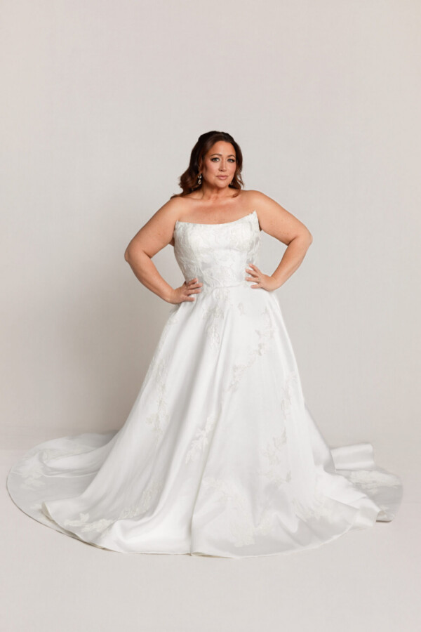 Bride wearing a one-shoulder white wedding gown with a full skirt and train, standing with hands on hips against a neutral background.