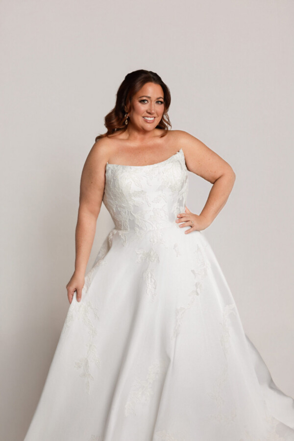 Close-up of a smiling bride in a one-shoulder white wedding gown with floral detailing, posing with one hand on her hip.