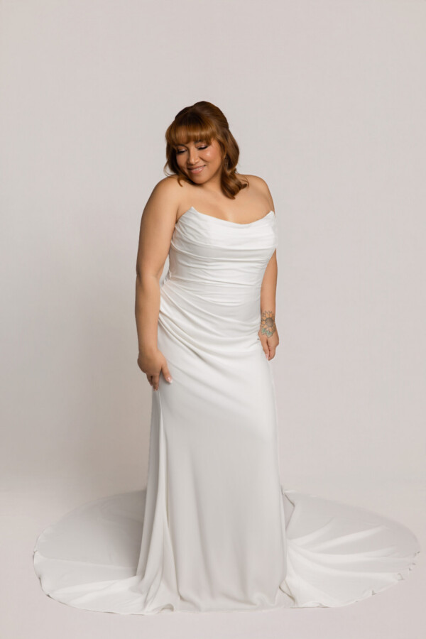 Bride wearing a draped one-shoulder white wedding gown with a fitted silhouette and slight train, posing against a neutral background.