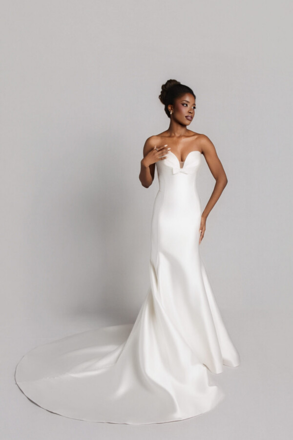 Bride wearing a strapless fitted white wedding gown with a sweetheart neckline and long train, posing against a neutral background.