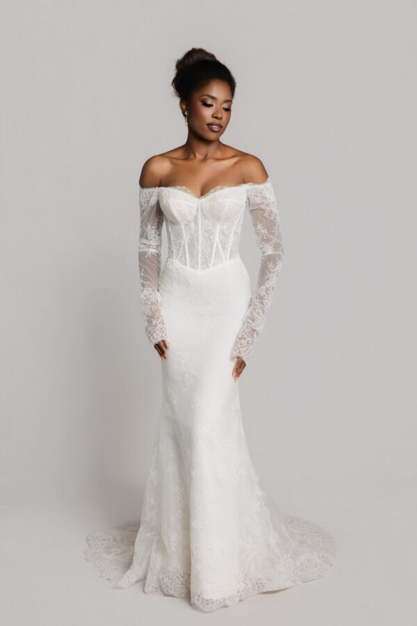 Bride wearing an off-the-shoulder lace wedding gown with long sheer sleeves and a fitted silhouette, standing against a neutral background.