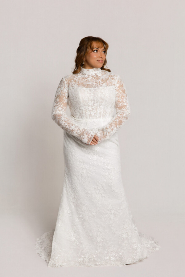 Bride wearing a high-neck long-sleeve lace wedding gown with a fitted silhouette, standing with hands clasped against a neutral background.