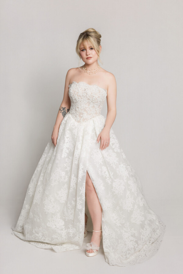 Bride wearing a strapless lace wedding gown with a fitted bodice and full skirt with a high slit, posing against a neutral background.
