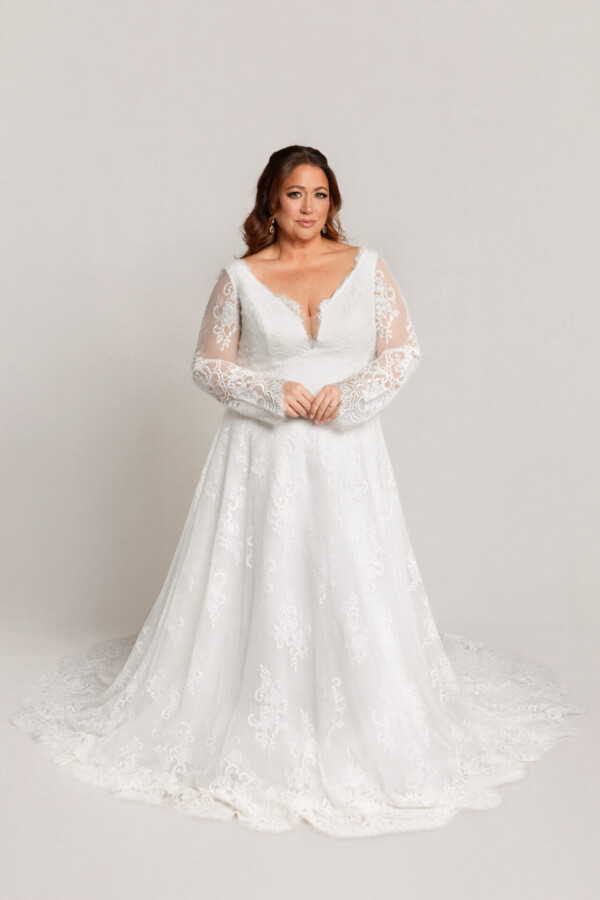Bride wearing a long-sleeve lace wedding gown with a V-neckline and full skirt, standing with hands clasped against a neutral background.