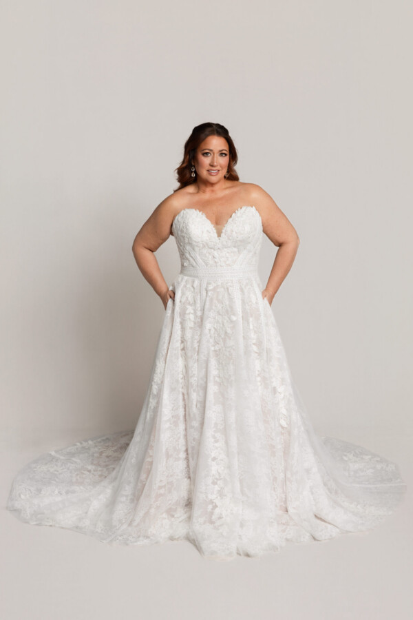 Bride wearing a strapless lace wedding gown with a sweetheart neckline and full skirt, standing with hands on hips.