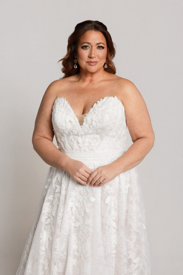 Close-up of a bride in a strapless lace wedding dress with a sweetheart neckline, standing with hands clasped against a neutral background.